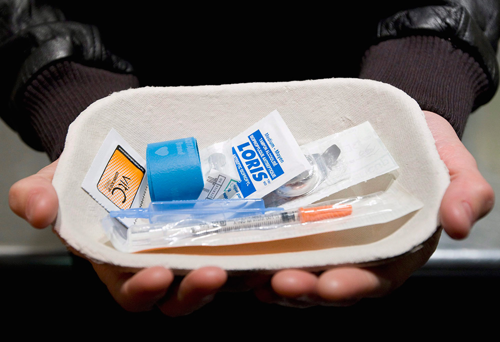 Two hands hold a small paper tray filled with safe injection supplies: an antiseptic wipe, syringe and tourniquet.
