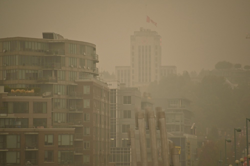A smoke-shrouded cityscape. The Vancouver City Hall building, with a Canadian flag flying atop it, can barely be made out through the haze.