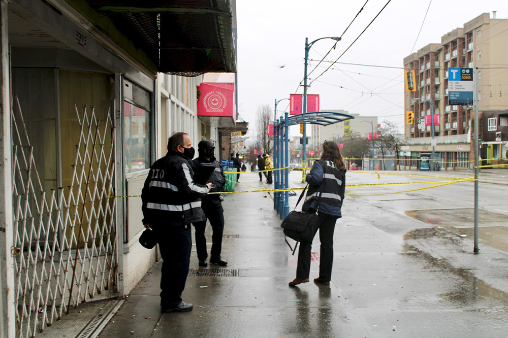 Two people with their backs to the camera and IIO written on their jackets stand on an urban sidewalk next to an area blocked off by police tape. Inside the police tape is a police officer in dark uniform with their face covered.