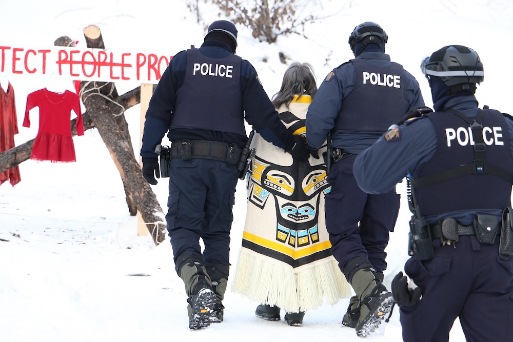 A woman wearing clothes with Indigenous markings is seen from behind being led away by several police in a snowy setting. 