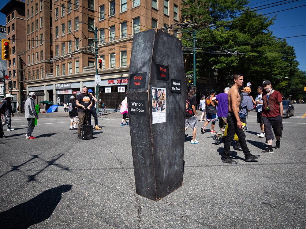 An empty black coffin stands upright at Main and Hastings Streets in Vancouver before a memorial march for victims of toxic drugs begin. The coffin has stickers saying “War on Drugs” and “War on Poor.”