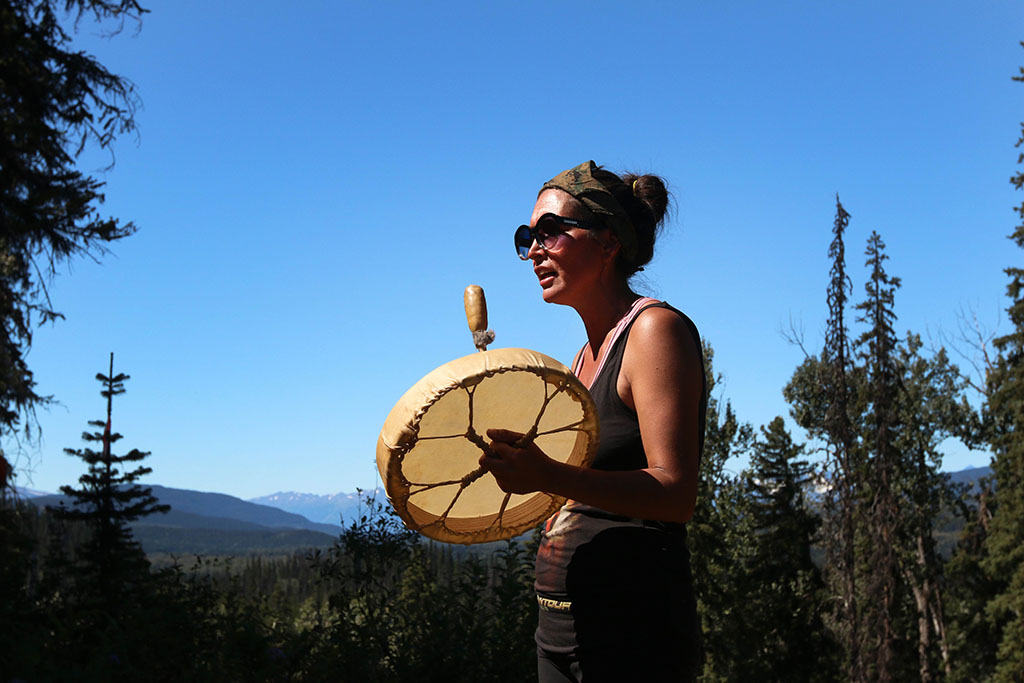 Gidimt'en Clan member Molly Wickham, Sleydo,' is holding a drum. She is wearing sunglasses and a tank top. Behind her is a forest, mountain range and blue sky.
