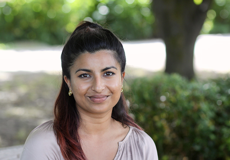 Anjali Appadurai, sitting outside in the shade in a light-coloured blouse, her black hair pulled back, smiles slightly at the camera.
