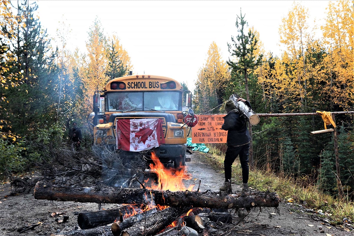 A roadblock preventing Coastal GasLink from accessing a site where it plans to drill under Wedzin Kwa, also known as the Morice River. The roadblock features a large campfire, a horizontal tree, and a school bus that bears an upside-down Canadian flag with red handprints on it. A person carries a log towards the school bus.