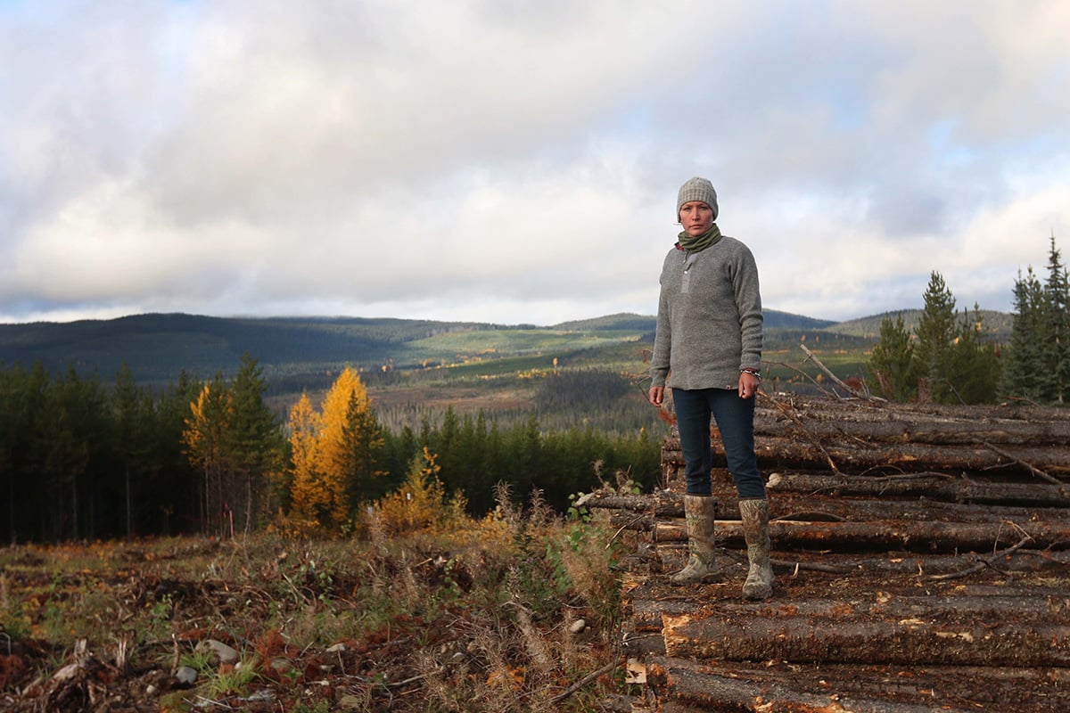 A woman in a grey sweater, toque and jeans stands in a muddy clearing beside a pile of newly cut logs. In the background are rolling hills and trees in fall colours.