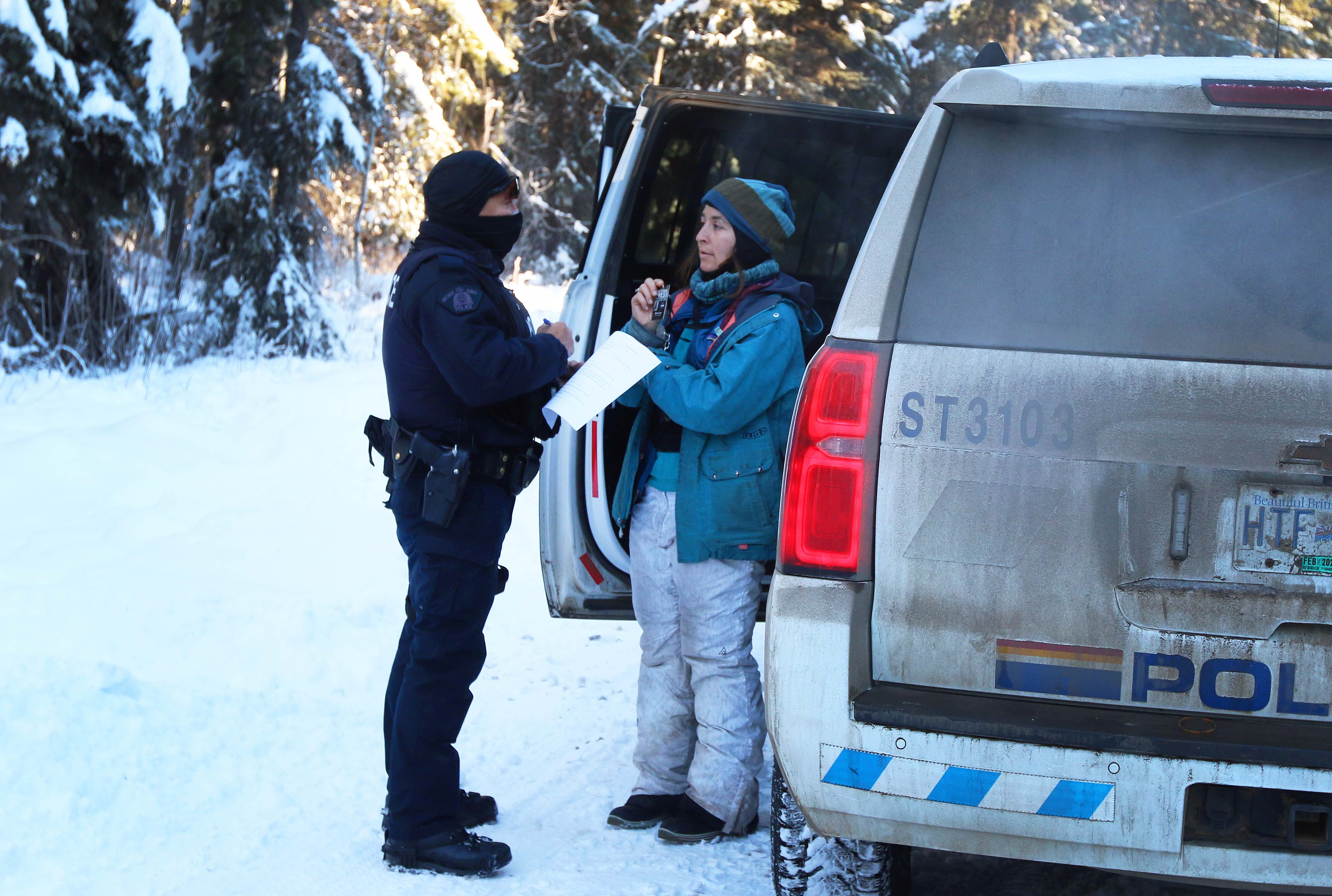 Two people stand facing each other next to a police vehicle. A police officer in uniform with their face covered takes notes as a woman, wearing a tuque and winter coat and pants, holds a card up in front of her. 