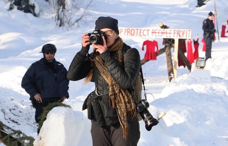 A woman with a long braid wearing a black down coat and black tuque holds up a camera. A second camera hangs over her shoulder. In the background, a police officer watcher here and red dresses are hanging.  