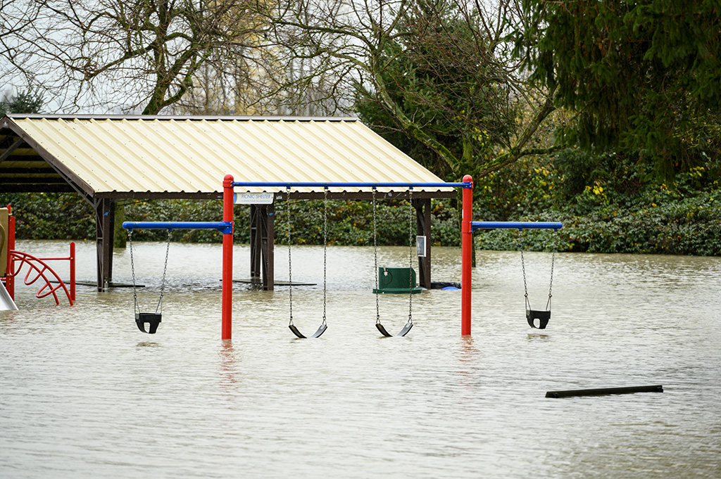 Grey water fills a playground up to the bottom of a red and white swing set in Abbotsford, B.C. Behind the swings is a covered picnic space, also flooded. Surrounding the playground is a stand of shrubbery and trees.