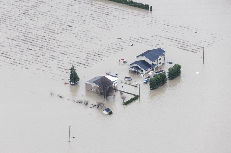 A farmhouse and adjacent farmland, seen from above, are surrounded by grey floodwaters. The cars outside the home are half covered.