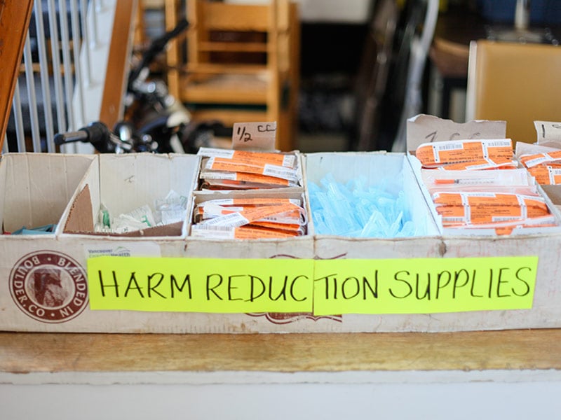 A table holds several boxes, including boxes of needles. The boxes are stuck together and affixed with a label reading, “Harm Reduction Supplies.”