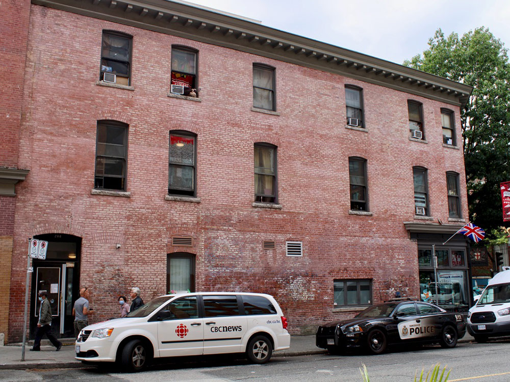 A three-storey, neglected red brick building with tall, narrow windows across the second and third floors. A CBC News van and police car are parked outside a black door.