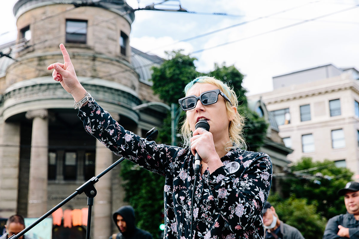 A person with sunglasses, dyed blond hair and a black button-down floral shirt raises a hand in the air while holding a microphone in the other. They are standing outside in front of an old concrete building with Corinthian columns on a cloudy day with a light blue sky.
