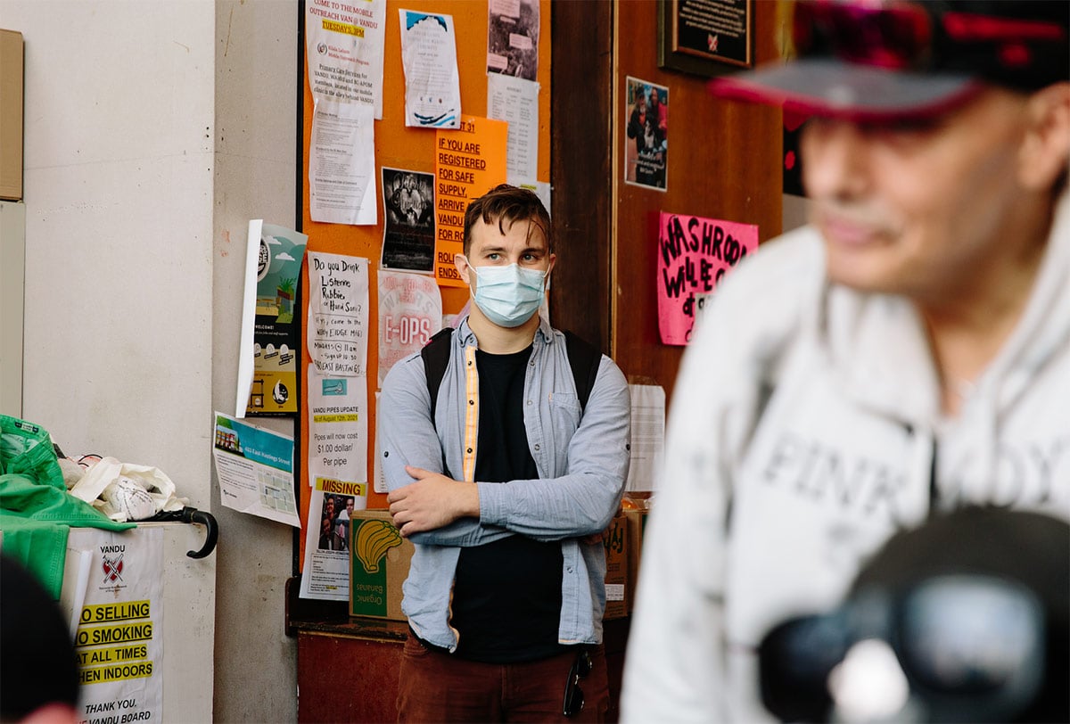 A white man wearing a blue button-down shirt over a black t-shirt, a backpack and a mask stands in the corner of a room beside a bulletin board covered with posters and announcements.