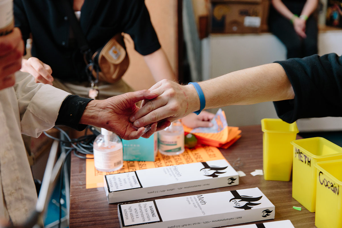 A hand with a blue bracelet reaches from the right side of the frame across a table to give something to a hand with a white sleeve reaching from the left side of the frame. They are reaching over a brown table laid with white rectangular boxes with minimalist black graphics on them. Behind them are small yellow sharps buckets with "meth" and "cocaine" in handwritten black marker on them.