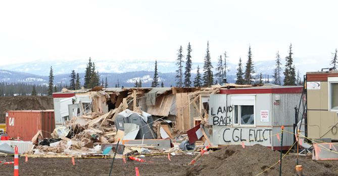Mobile site office trailers and a portapotty or two have been significantly damaged, with plywood and other debris scattered on the ground. Graffiti reads ‘LAND BACK, CGL EVICT.’ 