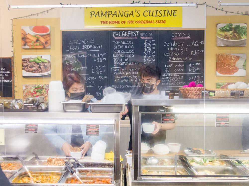 Edith Malang, left, and her son J, right, serve food from behind the counter at Pampanga’s Cuisine, a Filipino restaurant on Joyce Street in Vancouver. Both are wearing black medical masks. Edith is placing a full takeout container on the counter in front of her while J consults an order receipt as he packages a variety of meal items. In front of them, food is in stainless-steel warming trays. Behind them, menu items are handwritten on a chalkboard. 