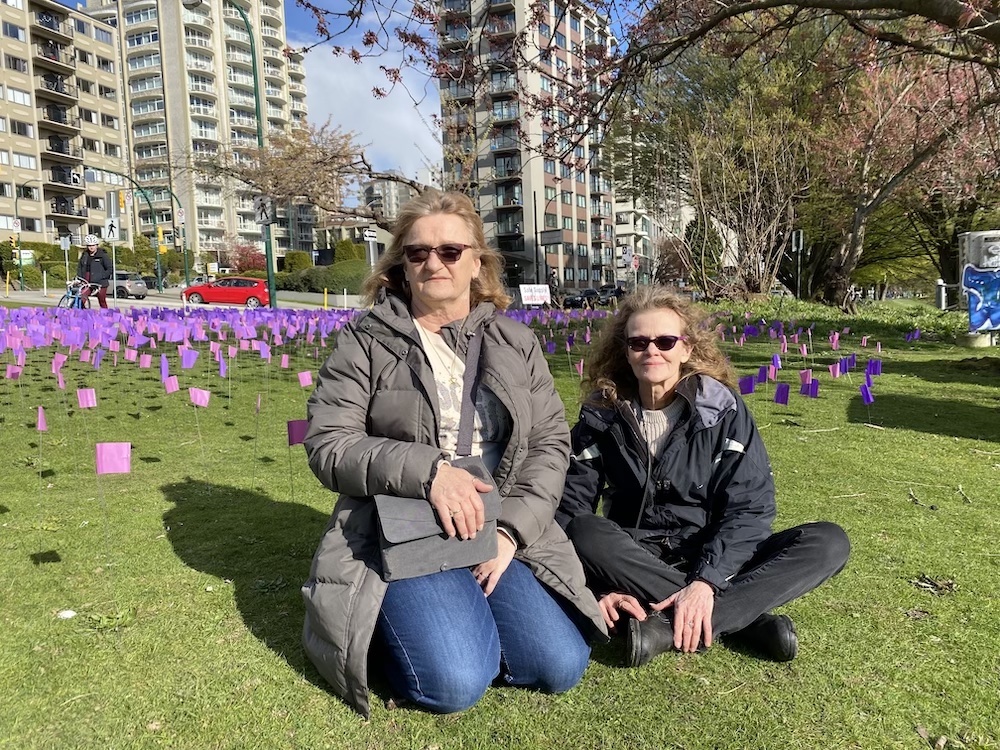 Debbie Lachapelle and her friend, Mona Murphy, sit on a grassy field in front of rows of purple flags. Cars and apartment building are in the background.