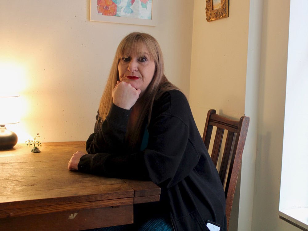 Tina Shaw sits and rests her face on her hand at a wooden table. A warm lamp on the table lights the room. Two framed pictures hang above her head.