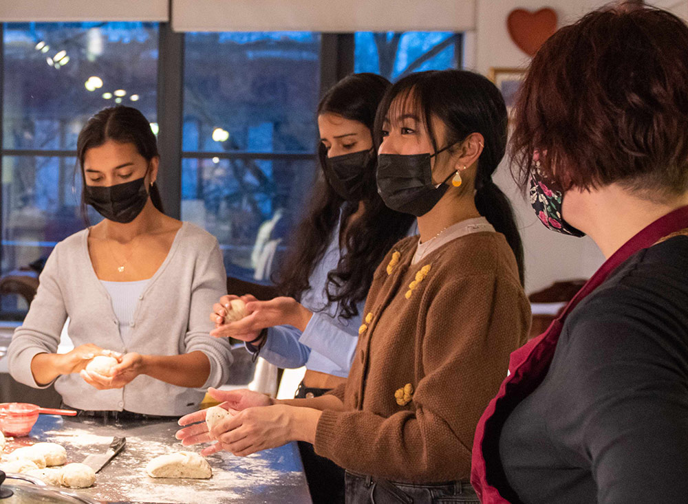 Four young people are wearing aprons and medical face masks as they gather around a kitchen counter, preparing food together. Three people are shaping pieces of dough with their hands. Another person looks on, hands on their hips.