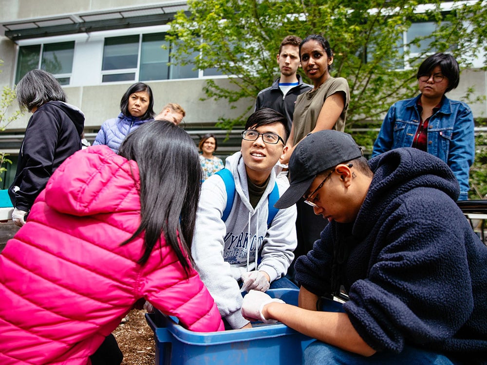 A group of people are gathered outdoors in a garden in front of a concrete building. In the foreground, three racialized people in their 20s are crouching and reaching into a tall blue plastic bin storing materials for planting and gardening. One person is looking up into the distance and smiling.