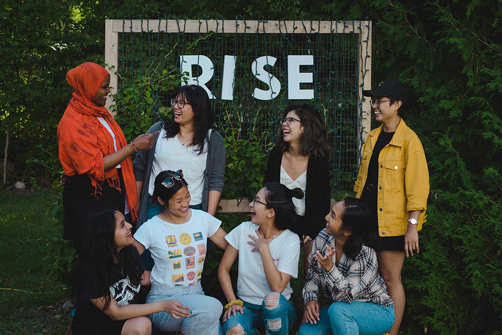 A group of eight young racialized people are gathered in front of a handmade sign that reads “Rise.” They are the founders of Vancouver’s Shades of Sustainability, which aims to create space for dialogue about environmental practices among BIPOC people. Members of the group are looking at each other, smiling and laughing.