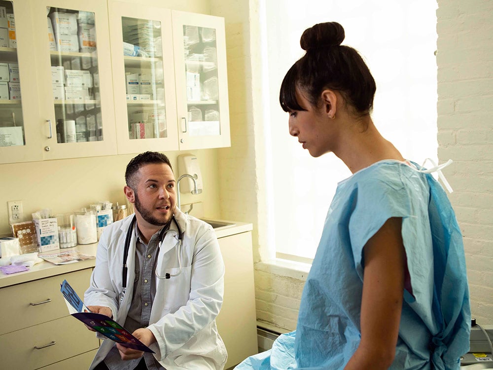 A transgender woman in a hospital gown speaking to her doctor, a transgender man, in an exam room.
