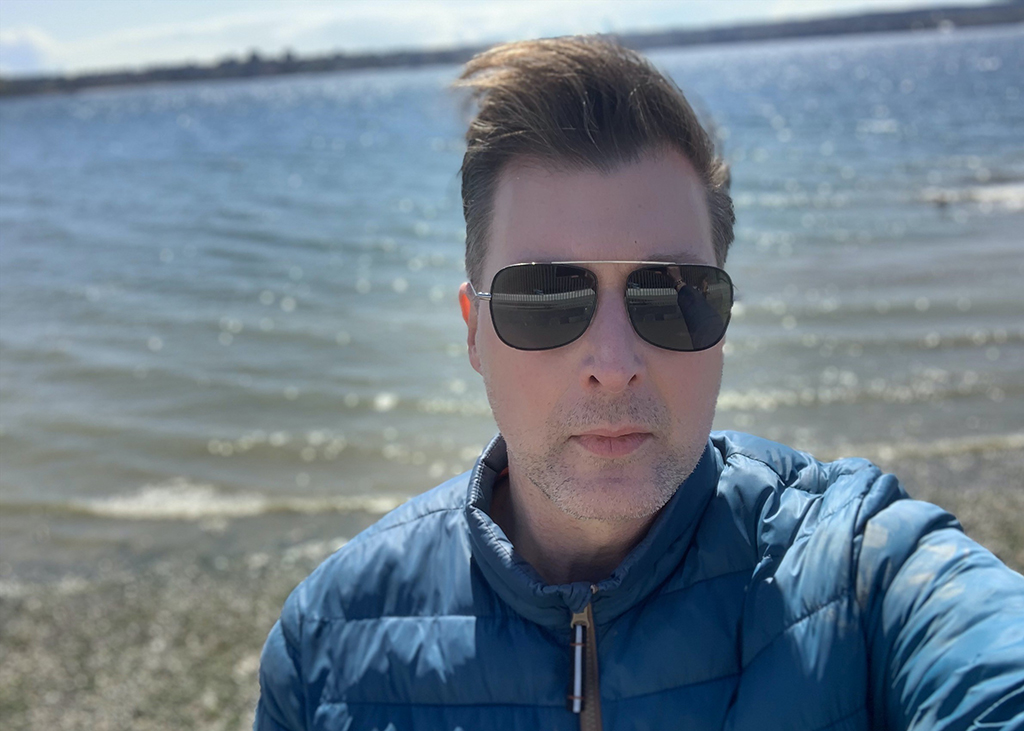 Darren Guy, in a blue jacket and sunglasses, stands in front of a body of water.