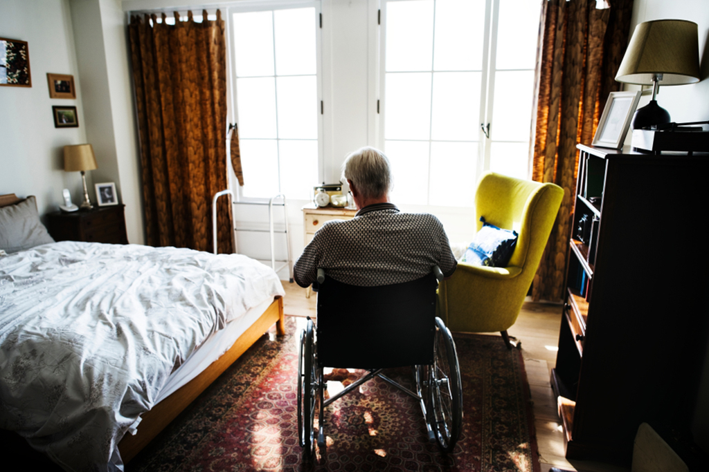 A man with white hair in a wheelchair is seen from behind sitting in bedroom with light from glass doors.