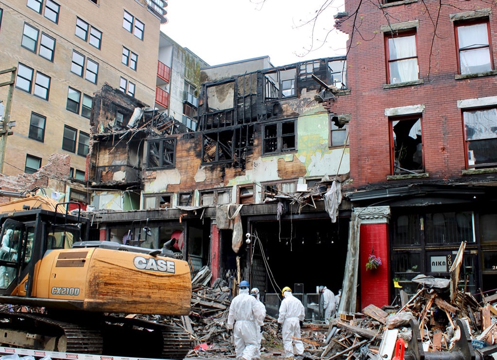 People in white hazmat suits in front of a heritage brick building which is partially demolished. Fire-blackened wood and walls can be seen in the partially destroyed building.