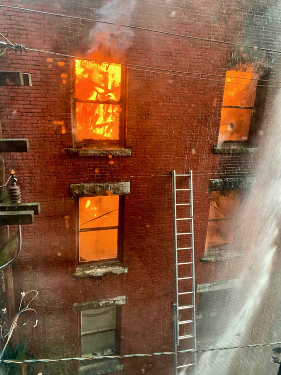 A red brick building, with flames visible through windows and a ladder propped against the wall.