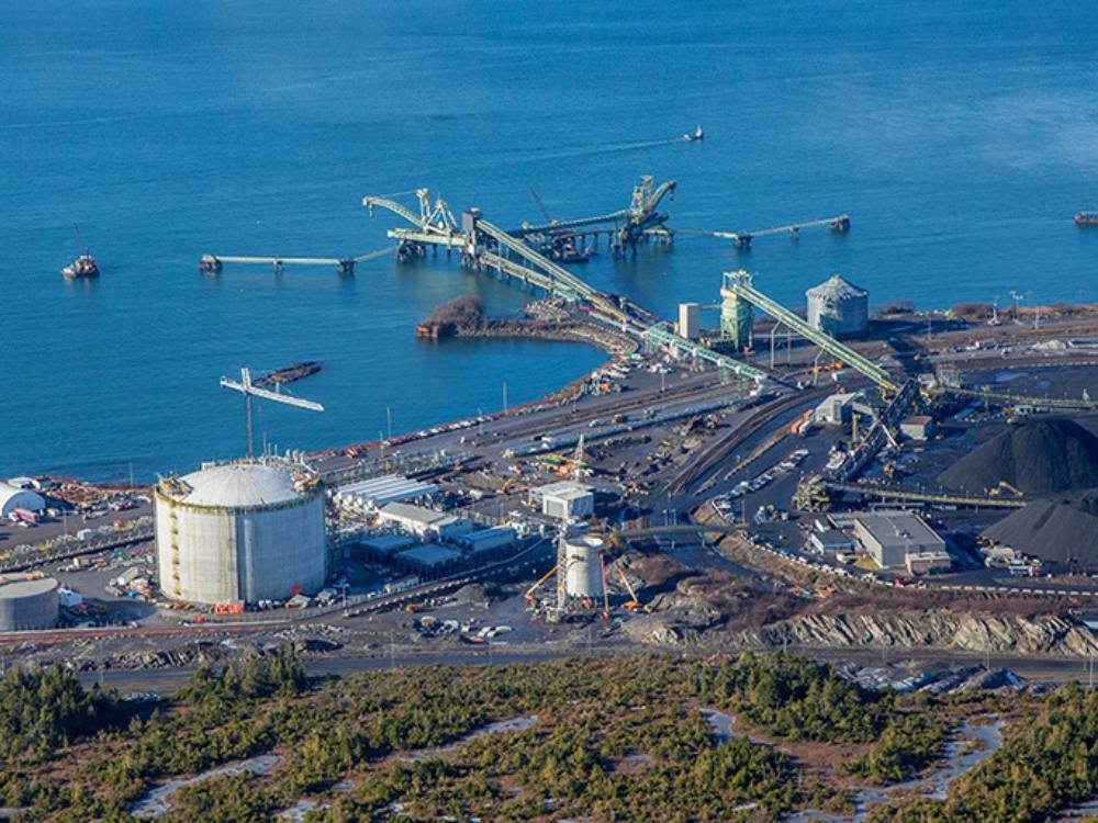 A large oceanfront scene shot from above, with giant piles of coal and equipment on the right and a large white concrete silo with a domed roof on the left.