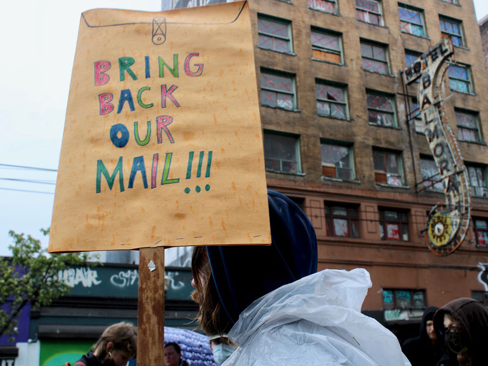 A man wearing a white plastic rain poncho holds a protest sign that says "bring back our mail." The Balmoral Hotel can be seen in the background.