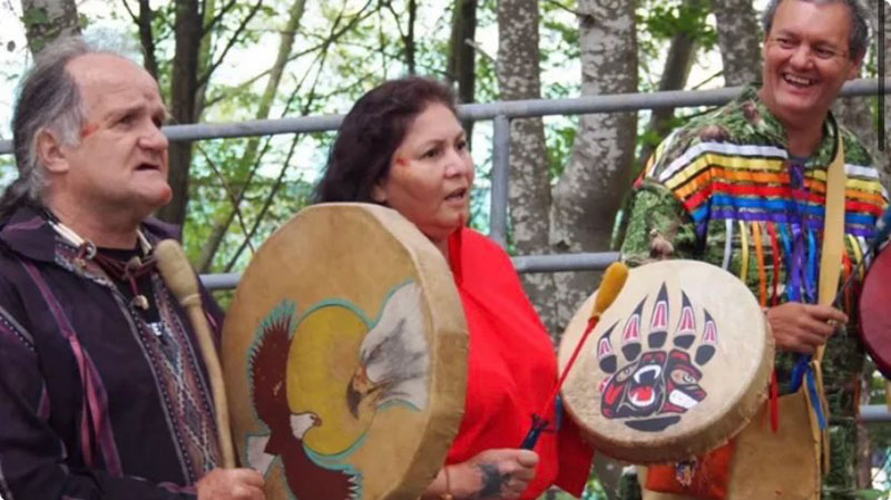 Three Indigenous people are drumming and looking at something beyond the camera. Black Bear Warrior is on the left and has an eagle painted on his drum. Beside him, a woman in red has a bear print on her drum. The pair are serious. A third man, whose drum is out of sight, is laughing. 