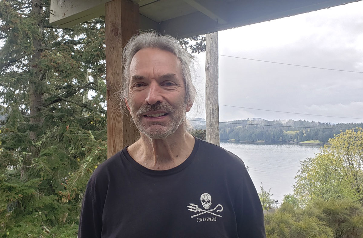Errol Povah's grey hair is pulled back in a low ponytail. Povah wears the iconic black Sea Shepherd Conservation Society T-shirt with a small skull and cross bones over the heart. Povah grins at the camera. In the background there are rolling hills and the waters of the Salish Sea.