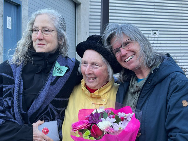 Three women with grey hair stand, arms around one another, grinning as they pose for the camera. Ruth Walmsley (left) has a flower print pinned on a green piece of cloth to her sweater, as a symbol for the Alouette Sisters. Catherine Hembling, in the centre, is wearing a bright yellow rain jacket.  