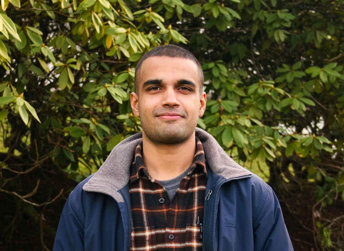 Zain Haq is a young man with buzzed hair, standing in front of a rhododendron bush. His jacket is open and he looks at ease as he smiles at the camera. 