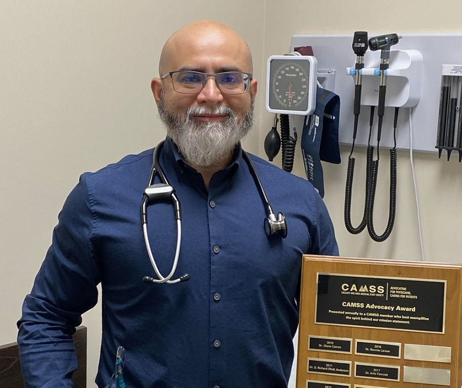 Dr. Mukarram Zaidi in a dark blue shirt holds his award in his office with stethoscopes in the background.