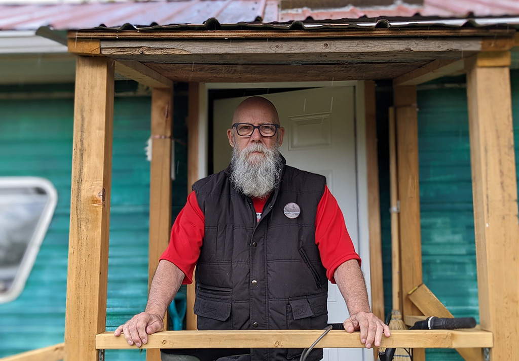Hunger striker Howard Breen, who is a bald, late-middle-aged white man with a long grey beard, stands staring at the camera on the porch of his cabin.