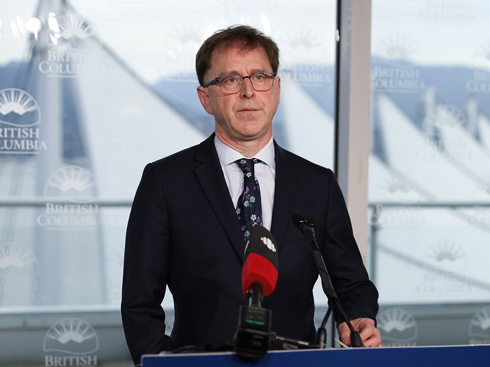 Adrian Dix, a man with brown hair and glasses, wearing a suit, stands in front of a podium.