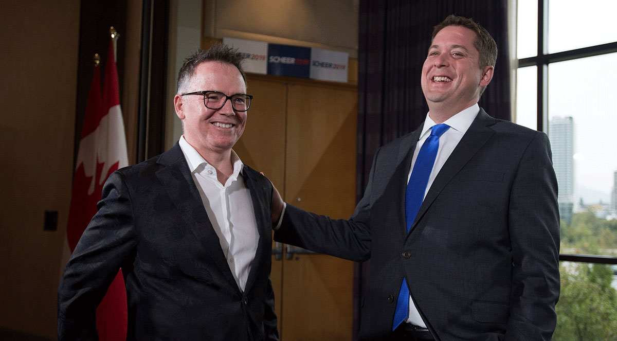 Two middle-aged white men in suits stand in a room laughing, the Canadian flag behind them.