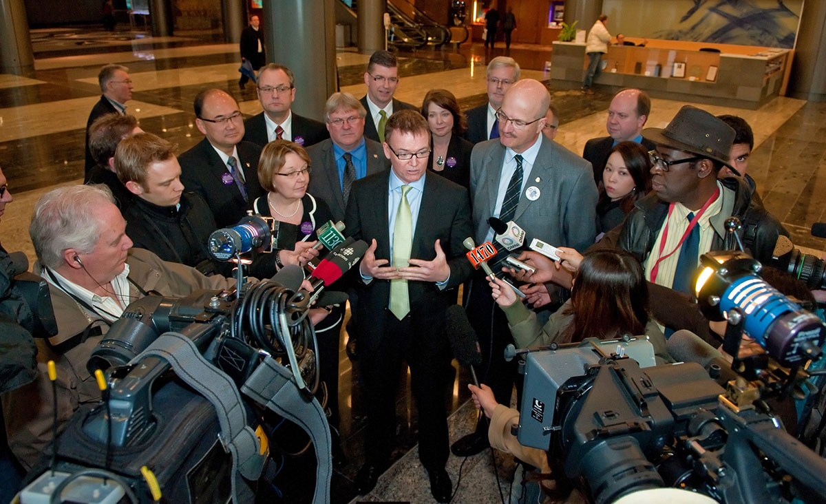 A man in a dark suit, gold tie and glasses — Kevin Falcon — stands flanked by BC Liberal politicians as he speaks to members of the press.