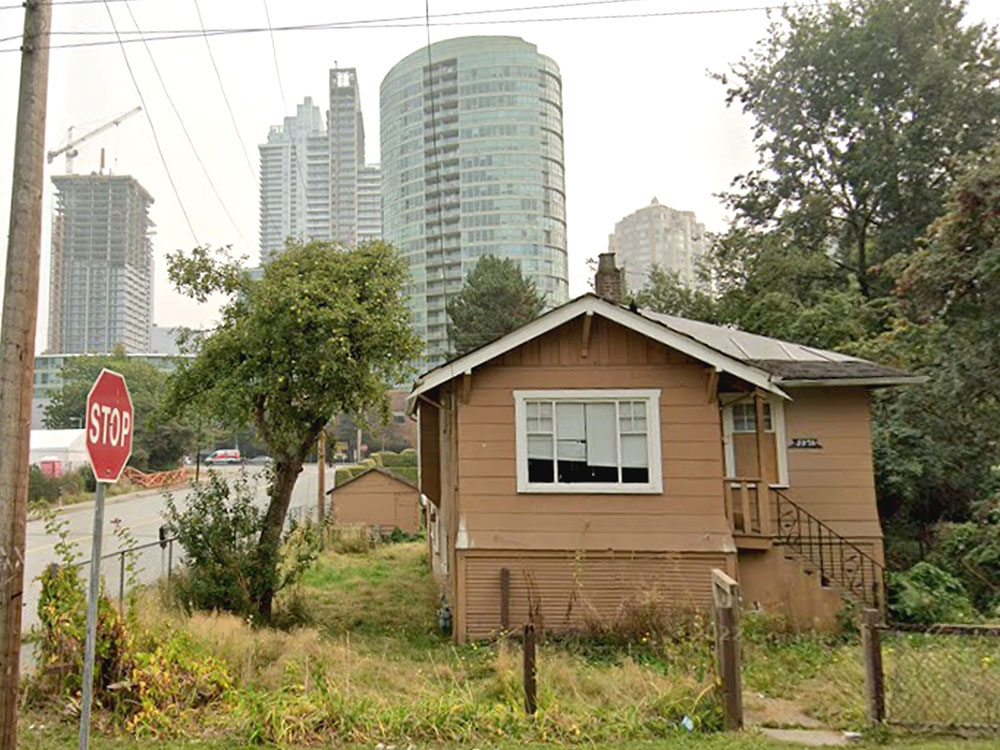 On the corner of a block is an old, brown, two storey-house with an overgrown lawn. On the left of the house is an apple tree with green fruit in its branches. Glassy high-rises tower over the house from behind.
