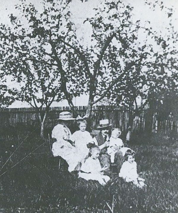 A faded black and white photograph of a mother and father, wearing hats, sitting in the grass with their four young children. Behind them are three leafy apple trees.