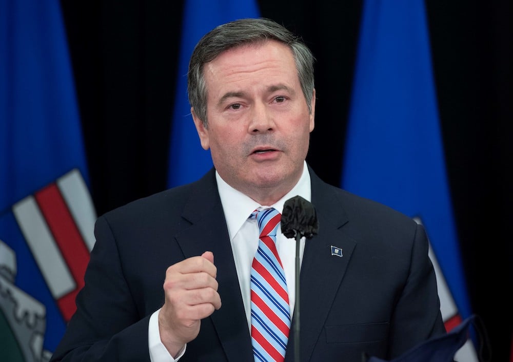 Jason Kenney, a middle-aged white man with dark hair, a blue and red striped tie and blue suit with a pin on his lapel speaks from a podium.