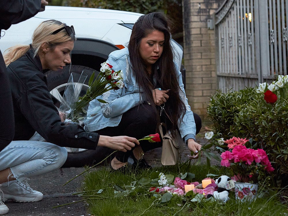 Two women kneel outside a large, gated brick home in front of a white sports utility vehicle, placing flowers at a shrine where more flowers and a stuffed panda sit.
