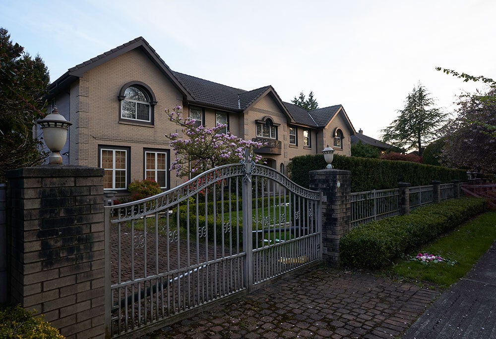 A large residence with sand-coloured bricks stands behind a locked white metal gate leading to the driveway.