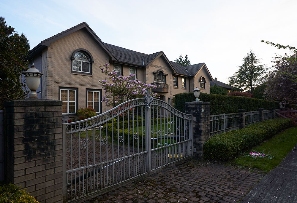 A large residence with sand-coloured bricks stands behind a locked white metal gate leading to the driveway.