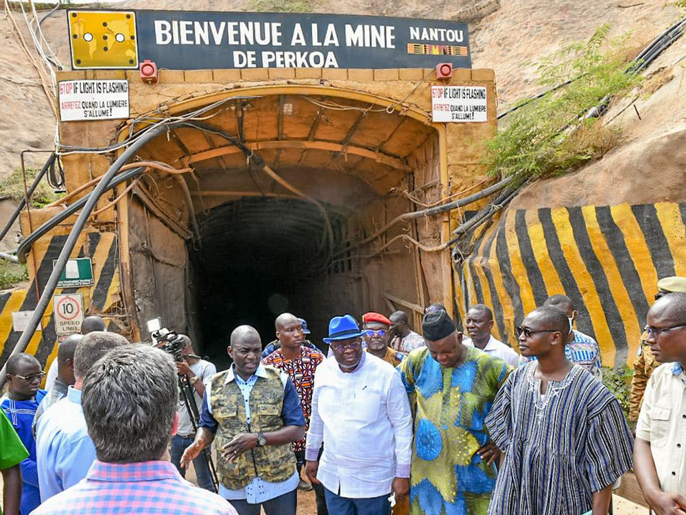 A group of about 15 men, some in traditional clothes of Burkina Faso, stand outside the entrance to a mine. Signs say “Bienvenue a la mine de Perkoa” and set out safety measures.