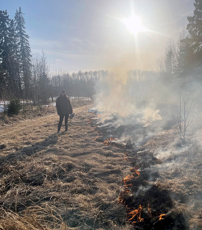 A silhouetted figure of woman in a black coat with a bright sun shrouded in smoky haze, with a long row of charred ground and small red flames receding from the foreground.