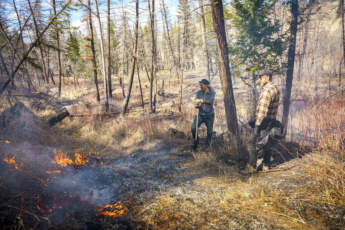 Two men, Russell Myers Ross and William Nikolakis, stand watching a patch of wild grass that is burning.